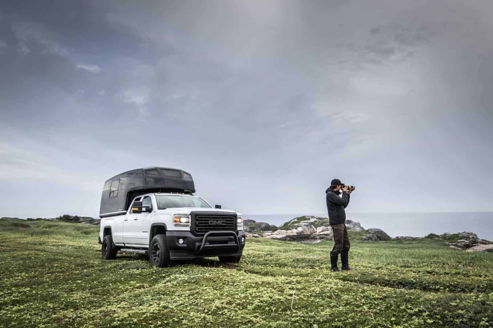 Un homme prenant des photos avec un appareil photo professionnel près d'un camion GMC blanc garé sur un terrain herbeux avec des rochers en arrière-plan sous un ciel nuageux.