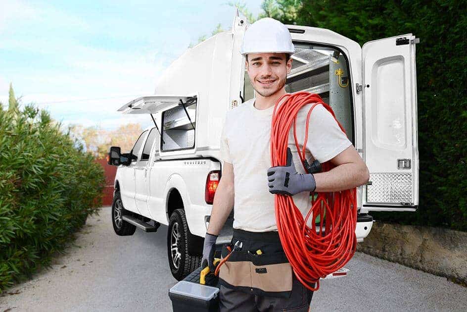 Électricien souriant avec casque blanc tenant un câble rouge et une boîte à outils devant une camionnette blanche ouverte