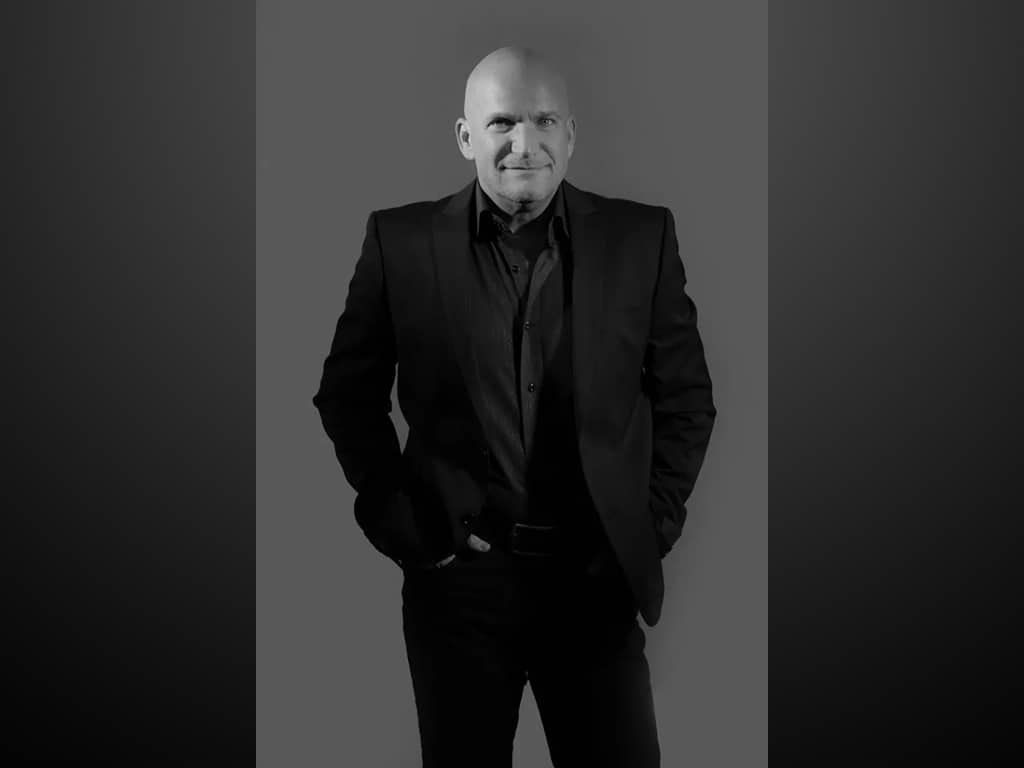 Bald man in a dark suit standing with hands in pockets against a dark background