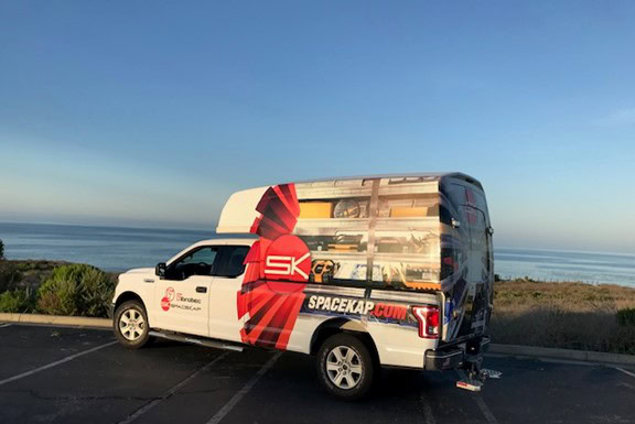 Camionnette Spacekap blanche avec un design rouge et noir stationnée près de la mer sous un ciel dégagé.