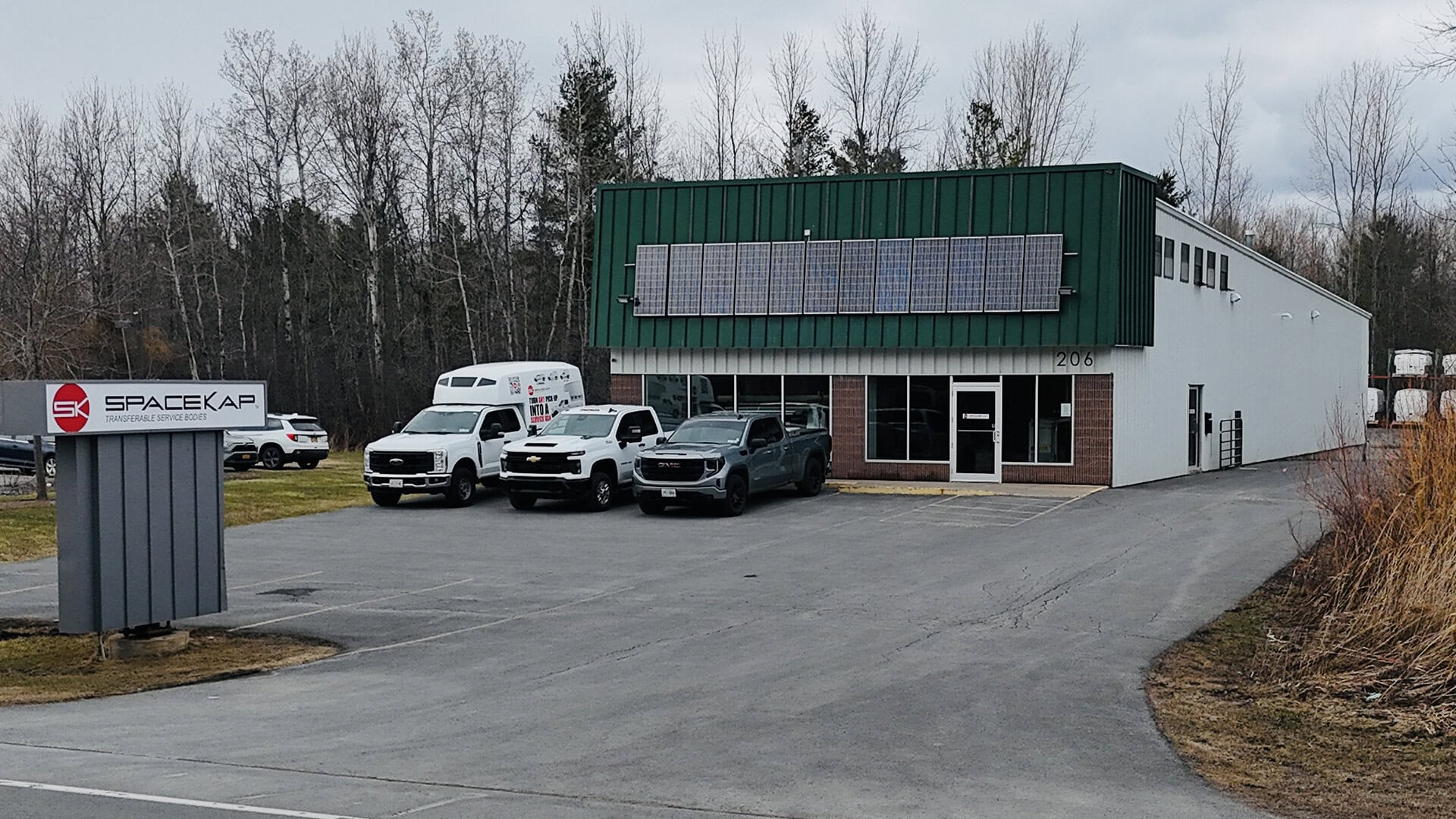 Commercial building with green upper facade and solar panels, trucks parked outside on cloudy day